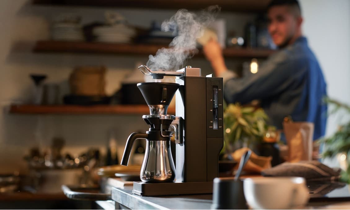 湯気の立つBALMUDAのコーヒーメーカー。奥のキッチンに男性。