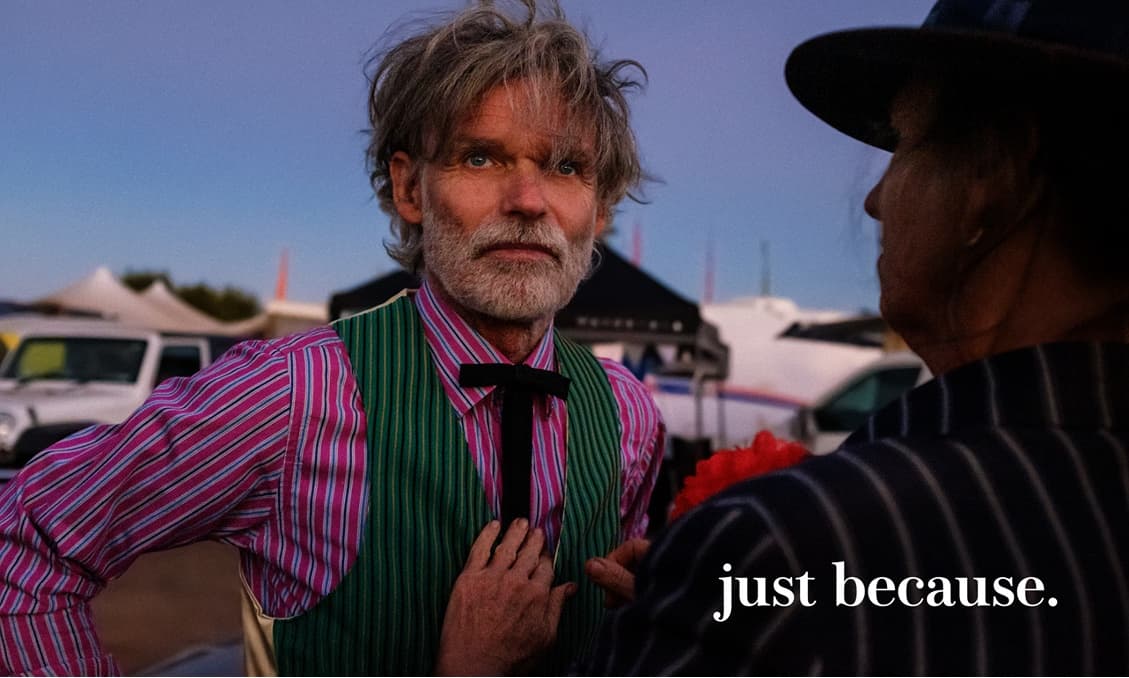 夕暮れの野外で、ストライプのシャツに緑のベストを着た白髪の男性。右下に「just because.」の文字。